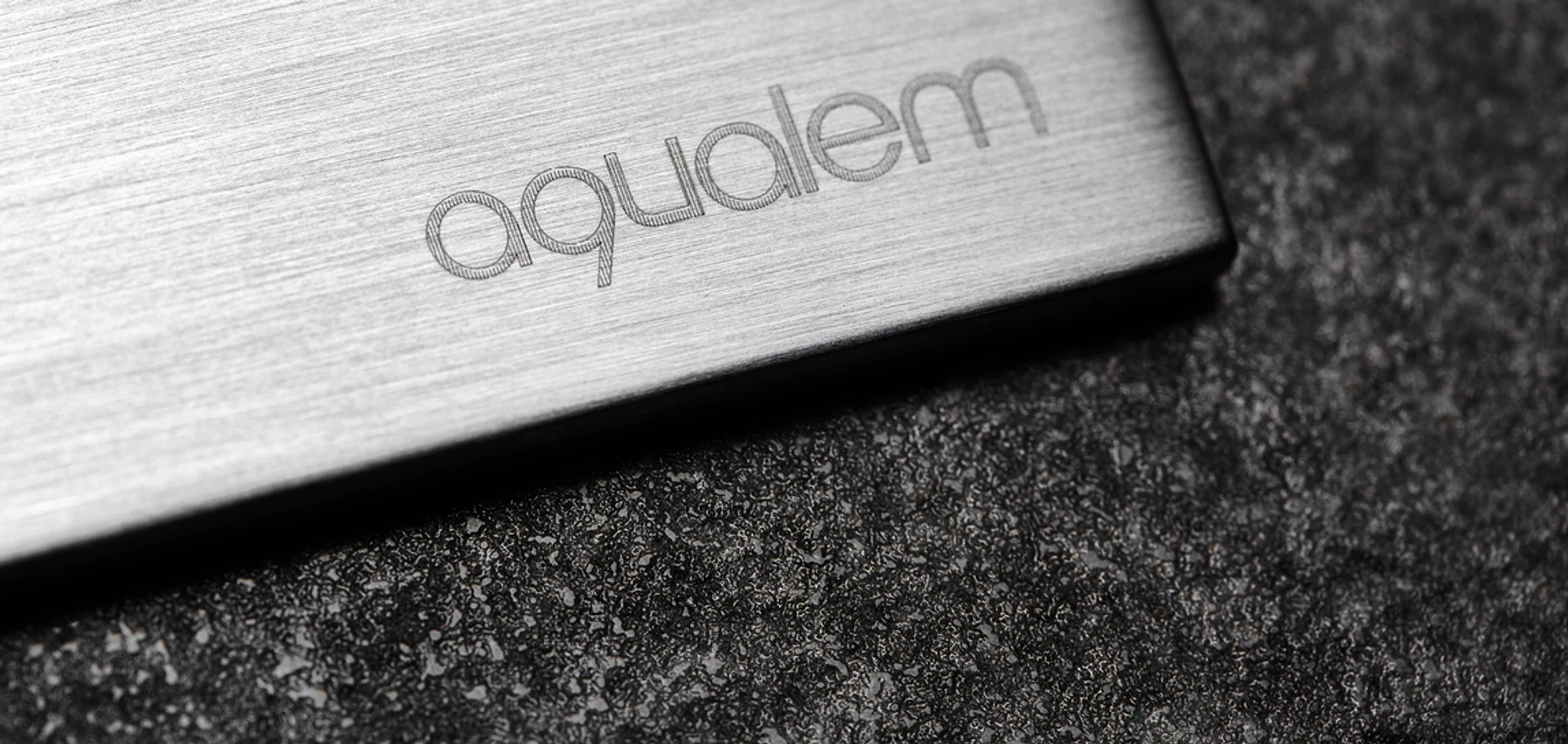 Brushed-metal Aqualem component engraved with the Aqualem wordmark, resting on dark stone.